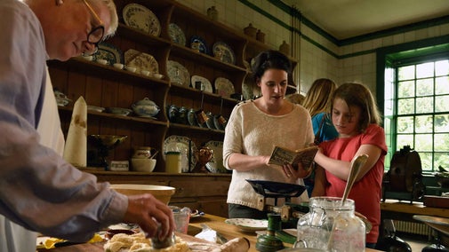 Visitors in the Kitchen at Wallington, Northumberland, look at items on the table, while a volunteer is baking